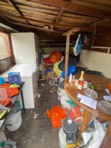 Interior of cluttered wooden shed containing white appliances, bags and household items requiring clearance